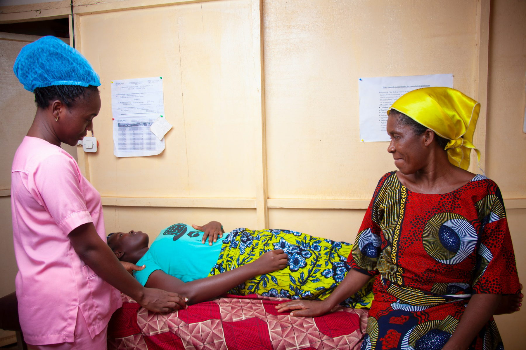 A midwife in a pink uniform and surgical cap examines a woman resting on a bed in a rural clinic room, while an older woman in a red print dress sits beside her.