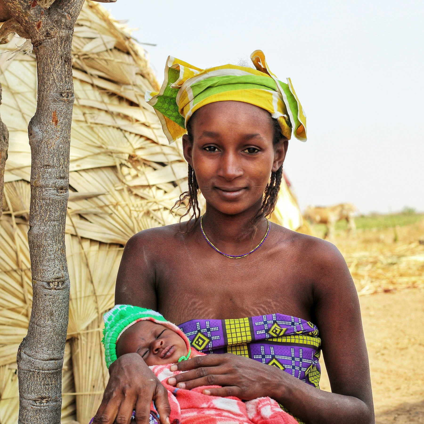 A young mother in a green and yellow headwrap holds her sleeping newborn, wrapped in a blanket, outside a thatched shelter in bright daylight.