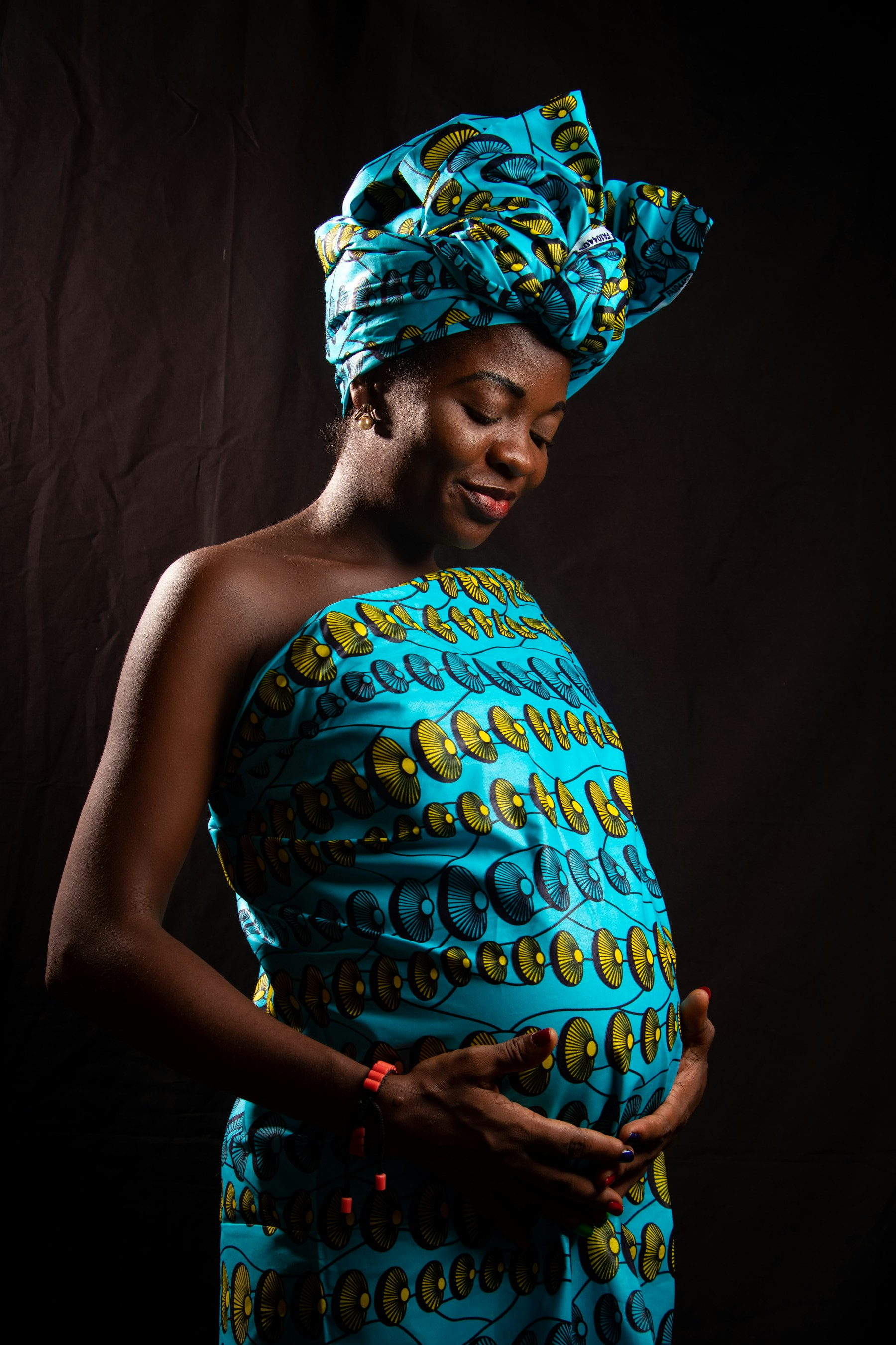 A pregnant woman in a turquoise and gold wax print wrap and matching headwrap rests both hands on her belly, looking down calmly against a dark background.